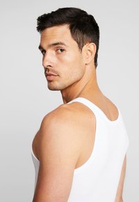White tank top made of cotton, featuring a snug fit, wide straps, and a rounded neckline. Model is looking over his shoulder.