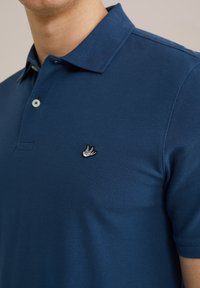 Temno modra polo majica iz teksturirane tkanine, z ovratnikom, dvema gumboma in majhnim vezenim ptico logotipom na levi strani prsnega dela.