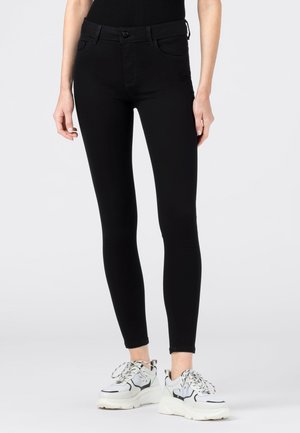 Pantalon noir slim porté avec des baskets blanches et noires épaisses sur une personne debout sur un fond uni.