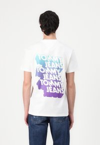 Hvid t-shirt med korte ærmer, prydet med overlappende "TOMMY JEANS" tekst i aqua og lilla gradient. Bagsiden viser en afslappet pasform og rund hals.