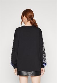 Svart långärmad tröja med avslappnad passform, med vita grafikdesigner på ärmarna och blå detaljer vid mudden.