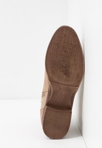 Semelle de chaussure marron avec motifs ondulés et nervurés, marquée de la taille 37, reposant verticalement contre un mur d’angle blanc.