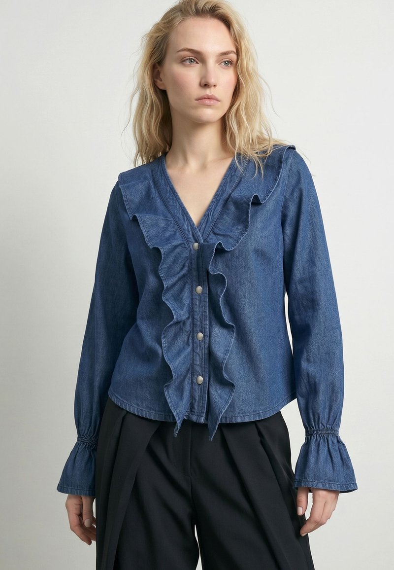 Femme blonde portant une blouse en denim bleu avec des volants sur le devant et une fermeture par boutons, associée à un pantalon noir ample sur un fond uni.