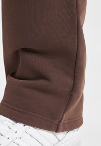 Pantalons de jogging marron en mélange de coton lisse, avec une coupe fuselée et des poignets élastiques, associés à des chaussures de sport blanches.