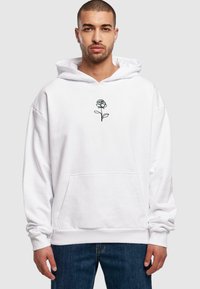 Weißer Kapuzenpullover mit einem schwarzen, liniengezeichneten Rosenmotiv auf der Brust, mit einer Fronttasche und gerippten Bündchen. Tragend zu blauen Jeans.
