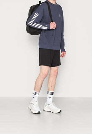 Felpa navy con strisce bianche, pantaloni corti neri, scarpe da ginnastica bianche e calzini a righe, abbinati a uno zaino nero. Look atletico semplice.