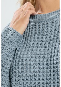 Jasnoniebieski sweter dziany z teksturowanym wzorem, okrągłym dekoltem i ściągaczami. Zbliżenie pokazujące rękę trzymającą dekolt.
