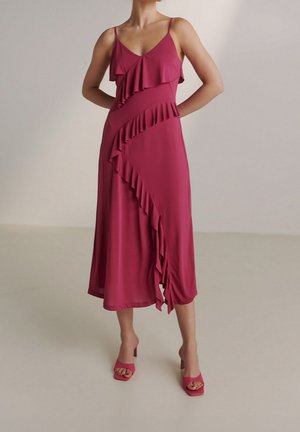 Femme portant une robe midi sans manches couleur magenta avec des volants diagonaux et des sandales à talons assorties, debout devant un fond uni.