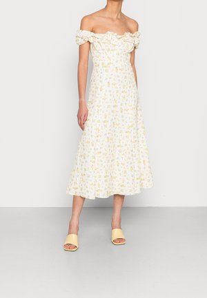 Nainen pukeutuneena valkoiseen, olkapäät paljastavaan midimekkoon, jossa on keltainen kukkakuvio ja röyhelökaulus. Mekon kanssa on yhdistetty keltaiset avonaiset liukusandalit.