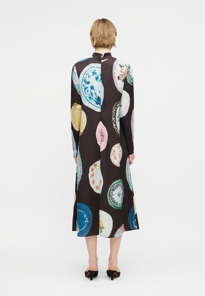 Άτομο που φοράει μακρύ, σκούρο φόρεμα με πολύχρωμα κυκλικά σχέδια πιάτων, στέκεται με την πλάτη γυρισμένη, φοράει μαύρα παπούτσια με τακούνι σε λευκό φόντο.