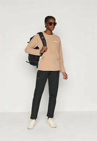 Maglia beige a maniche lunghe con logo bianco, pantaloni neri con accenti laterali, scarpe sportive bianche e uno zaino nero. Occhiali da sole scuri indossati.