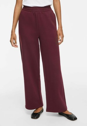 Femme portant un pantalon bordeaux taille haute à jambes larges, un haut blanc et des chaussures plates noires, debout sur un fond uni.