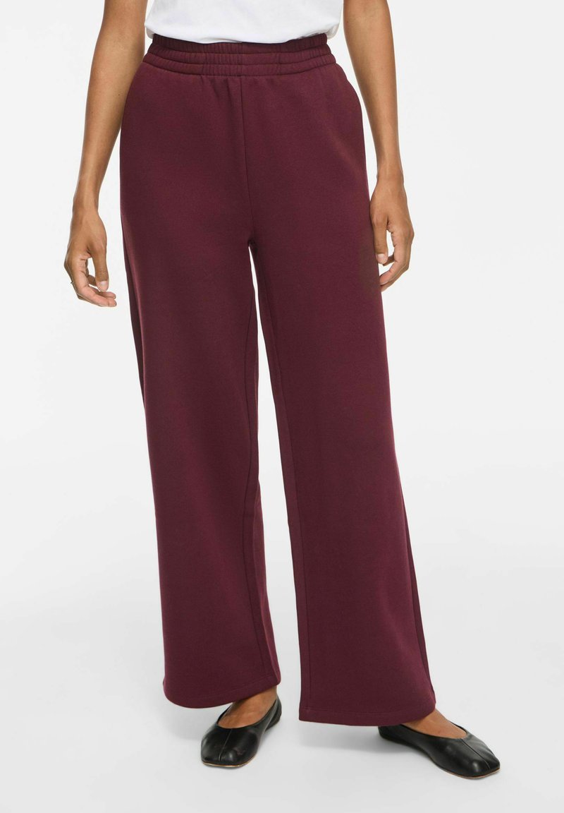 Femme portant un pantalon bordeaux taille haute à jambes larges, un haut blanc et des chaussures plates noires, debout sur un fond uni.