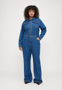 Combinaison en denim bleu moyen, avec des manches longues, un devant boutonné, deux poches poitrine, des jambes larges et une taille ceinturée pour plus de structure.