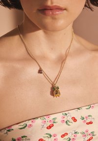 Närbild på en person som bär ett guldsmycke med en baggeformad hängsmycke med gröna och rosa ädelstenar, iklädd blommigt tyg.