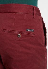 I pantaloni di cotone color bordeaux presentano una superficie testurizzata, una chiusura con bottone in metallo e un'etichetta applicata con il marchio "SCOTCH & SODA" sulla tasca.