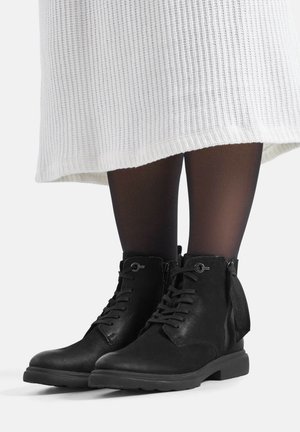 Czarne skórzane botki za kostkę z wiązaniem z przodu, zamkami po bokach i fakturowanym wykończeniem. Noszone z czarnymi rajstopami i białą prążkowaną spódnicą.