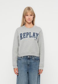 Šedá bavlnená mikina s potlačou "REPLAY" v námorníckej modrej na hrudi, s okrúhlym výstrihom a ribovanými manžetami, skombinovaná s modrými džínami.