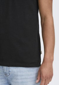 Camiseta de algodón negra con cuello redondo, mangas cortas y una pequeña etiqueta en el dobladillo. La tela tiene una textura suave.