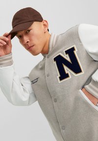 Grijze wollen varsityjack met witte leren mouwen, marineblauwe 'N' logo, geribbelde manchetten en knoopsluiting; gecombineerd met een bruine pet.