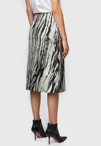 Femme portant une jupe midi à motifs abstraits noir et blanc, un haut blanc et des bottines à talons aiguilles noires pointues, se tenant sur le côté.
