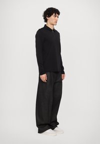 Svart langermet polo med glidelås, kombinert med vide mørke bukser og hvite sneakers. Stoffet ser glatt ut med minimal tekstur.