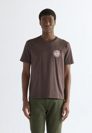 Jonge man met kort haar, wearing een donkerbruin T-shirt met een klein rond logo op de borst en olijfgroene broek, staand tegen een effen achtergrond.