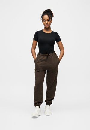 Femme debout les mains dans les poches, portant un haut noir ajusté à manches courtes, un pantalon de jogging marron et des baskets blanches sur fond blanc.