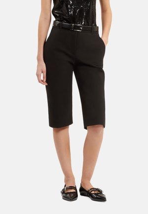Bermuda shorts neri su misura con una texture liscia, dotati di passanti per cintura e tasche laterali, abbinati a ballerine nere.