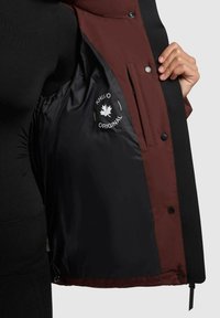 Una mano tiene aperto un giubbotto nero e marrone, mostrando la fodera interna nera con il logo bianco "Khujo Original" e un grafico di un acero.