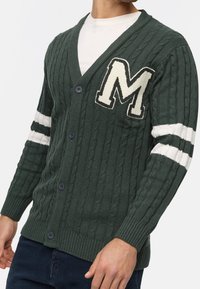Cardigan verde a maglia cable con accenti a strisce bianche e una grande "M" ricamata sul petto. Presenta cinque bottoni e polsini a coste.