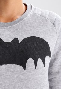 Felpa grigia con una grande silhouette di un pipistrello nero stampata sul petto e dettagli di cucitura visibili sulla spalla.