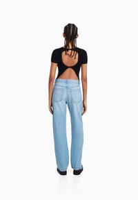 Mujer con cabello trenzado lleva una blusa negra de manga corta con la espalda descubierta y unos jeans rectos de tiro alto de color azul claro, de pie mirando hacia atrás.