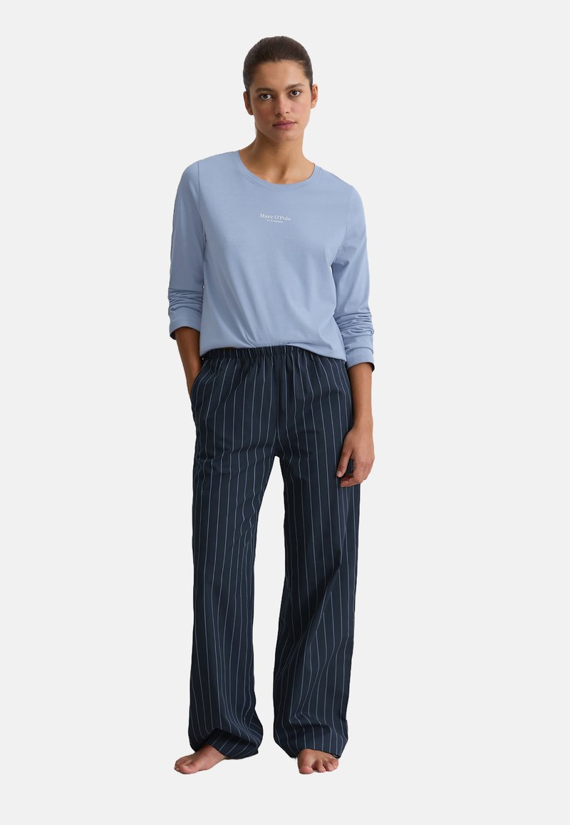 Vrouw die barefeet staat en een lichtblauw langemouwenshirt en een donkerblauw gestreepte wijde broek draagt met handen in de zakken.