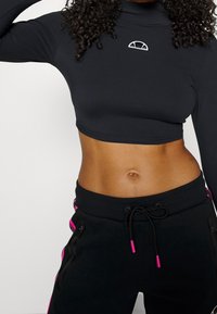 Musta lyhythihainen toppi, jossa on korkea kaula-aukko ja pieni graafinen logo rinnan yläpuolella. Yhdistetty mustiin housuihin, joissa on vaaleanpunaisia yksityiskohtia.