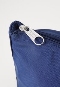Esquina de bolsa de nylon azul marino con superficie texturizada, que presenta un tirador de cremallera plateado con la inscripción "TOMMY JEANS". Primer plano del detalle de la cremallera.