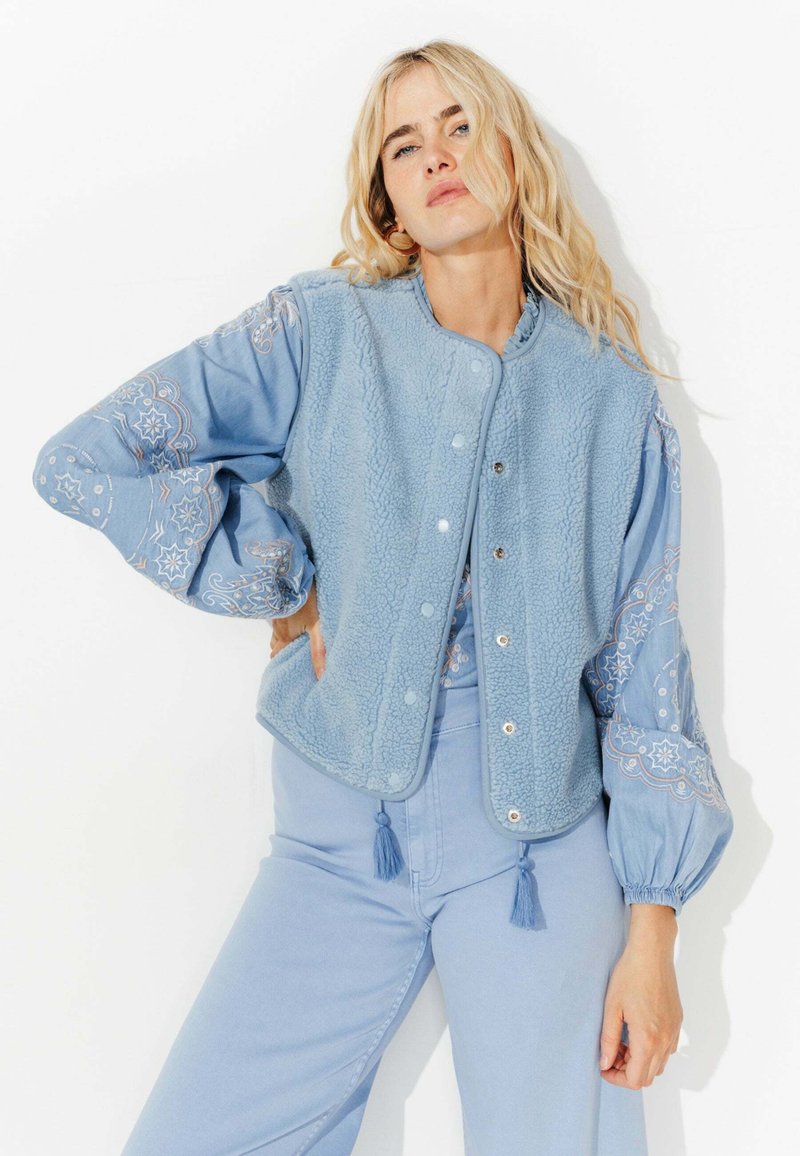 Veste bleu clair avec un corps en polaire texturé et des manches brodées, dotée de boutons-pression et d'accents à frange en bas.
