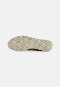 Suola di scarpa realizzata in materiale gommoso testurizzato, con un motivo a zigzag per migliorare la trazione. Il colore è principalmente bianco con accenti verdi ai lati.