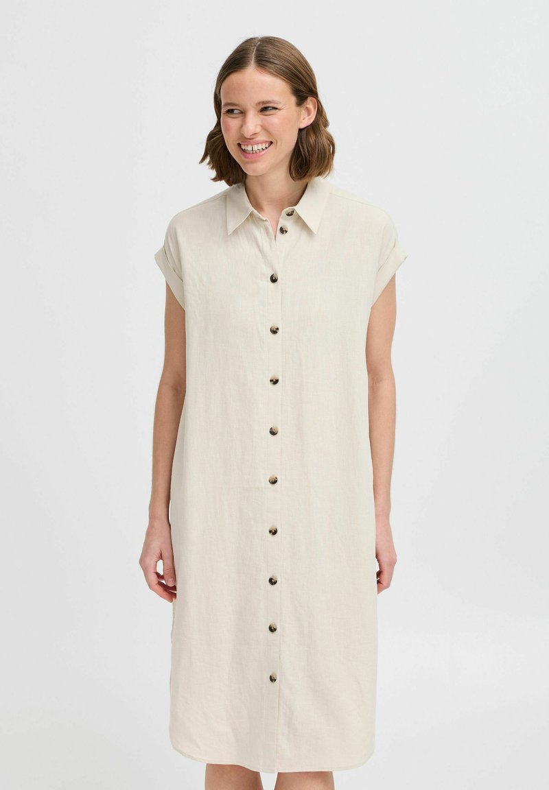 Abito camicia in lino color crema con bottone frontale, maniche corte arrotolate e una vestibilità comoda. Presenta un orlo dritto e una texture sottile.