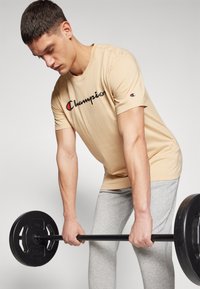 Beige Baumwoll-T-Shirt mit "Champion"-Logo, kombiniert mit hellgrauen Sporthosen. Person hebt eine Langhantel mit schwarzen Gewichten.