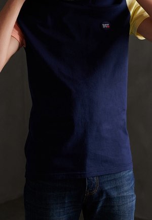 Person trägt ein dunkelblaues T-Shirt mit kurzen gelben und pfirsichfarbenen Ärmeln und blauen Jeans, mit einem kleinen Superdry-Logo auf der Brust.