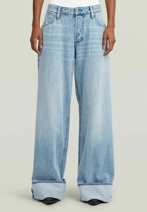 Person wearing light blue wide-leg jeans with rolled cuffs and black shoes, standing against a plain background.