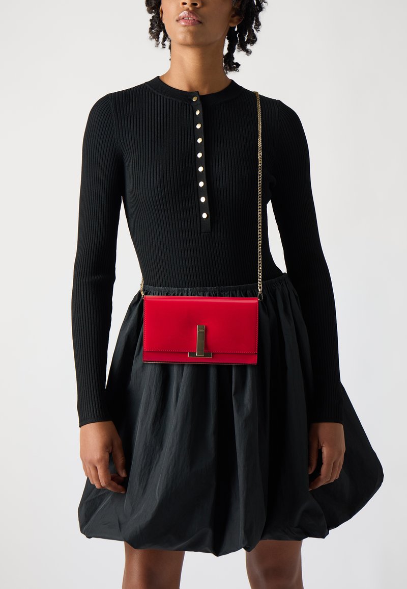 Rød skinncrossbody-veske med rektangulær form, utstyrt med en sølvfarget lås og en kjedeavtak som bæres over en svart ribbet topp.