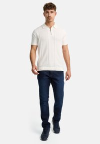 Camisa polo de hombre en crema con cremallera corta, tela texturizada, cuello de canalé, combinada con jeans de mezclilla oscura y zapatillas oscuras.