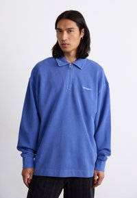 Blue zip-up sweatshirt with a collar, made of textured fabric. Features a small white logo on the chest and long, loose sleeves.
