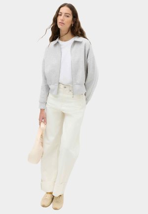 Vrouw met lichtgrijs jack met rits, witte blouse, off-white broek, beige schoenen, met een zachte beige handtas tegen een minimalistische achtergrond.