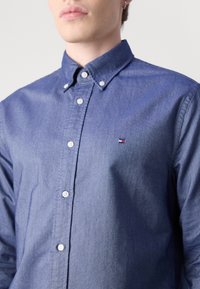 Un homme porte une chemise bleue à manches longues boutonnée avec un petit logo rouge, blanc et bleu sur la poitrine gauche et un col à boutons.
