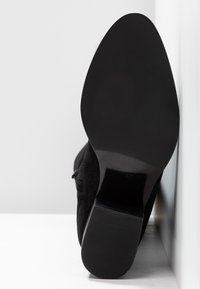 Bottine en daim noir avec un bout arrondi et un talon épais. La semelle est lisse et plate, avec une texture côtelée.