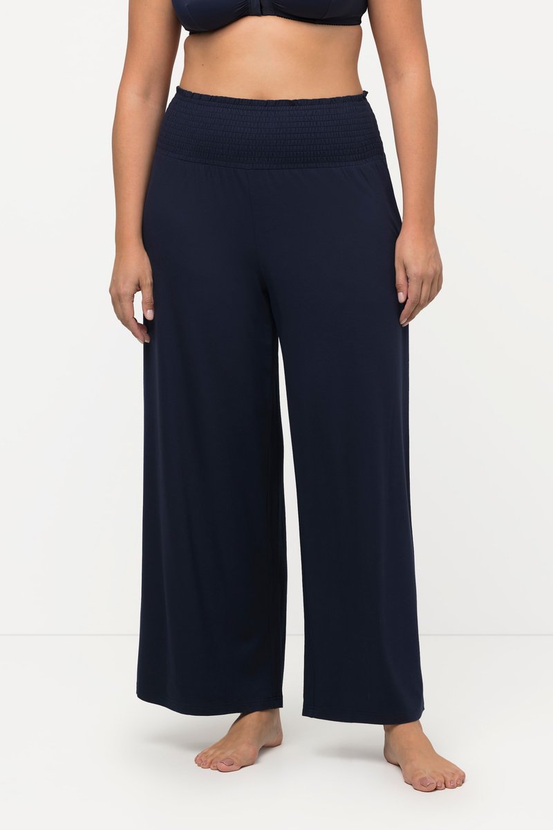 Donna che indossa pantaloni larghi blu navy scuro con una fascia elastica larga in vita, a piedi nudi, in piedi davanti a uno sfondo bianco semplice.