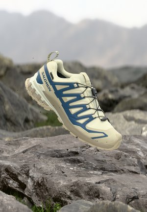 Letvægts beige trailrunningsko med blå accenter, tekstureret mesh-overside og en robust sål, der har elastiske snørebånd og en træktab.